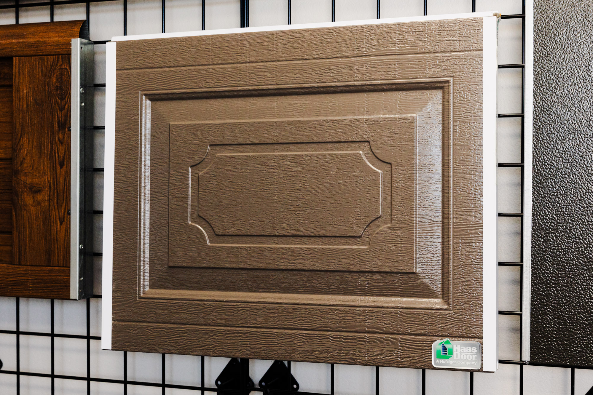 Brown raised-panel garage door sample displayed on a wall.